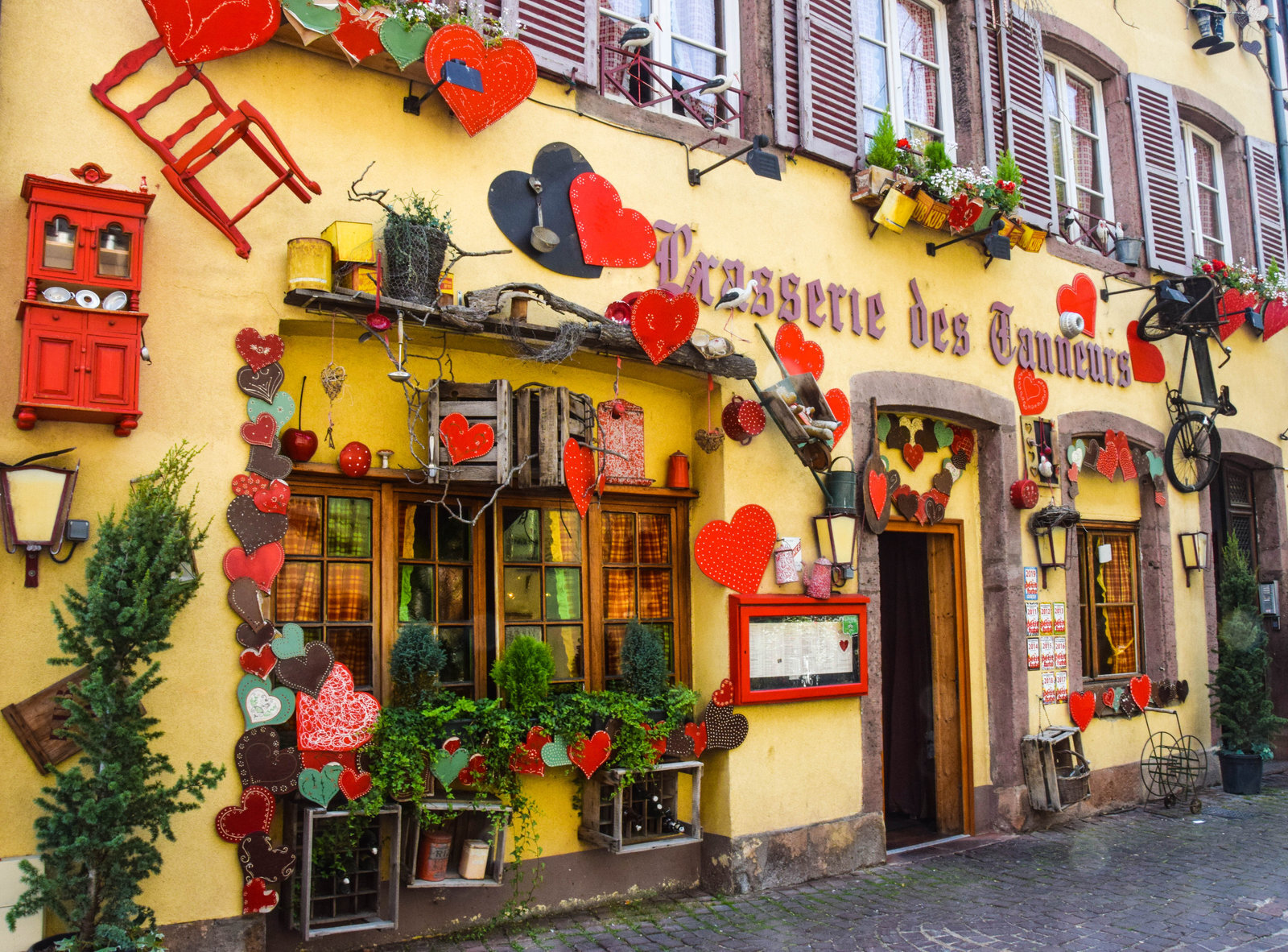 Façade jaune de la Brasserie des Tanneurs, colombages, cœurs alsaciens en bois rouge accrochés autour des fenêtres